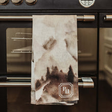 Load image into Gallery viewer, A beige kitchen towel with a dark brown abstract print and a monogrammed 'PH' logo, hanging from the handle of a black Belling double oven.