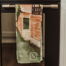 Load image into Gallery viewer, A kitchen towel featuring a printed architectural scene in muted green, terracotta, and cream tones, hanging from a metal handle on a black oven door.