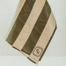 Load image into Gallery viewer, A hand with red nail polish holds up a striped kitchen towel or napkin with alternating olive green and cream vertical stripes, featuring a small embroidered monogram logo in the bottom corner.