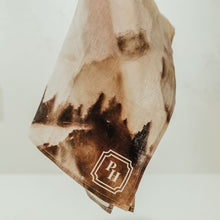 Load image into Gallery viewer, A hand with red nail polish holds up a lightweight, semi-sheer scarf featuring a brown and beige watercolor mountain pattern and a decorative 'PH' monogram logo.