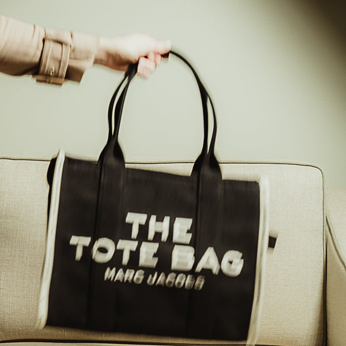 Black tote bag with 'The Tote Bag' and 'Marc Jacobs' text held by a person on a beige couch.