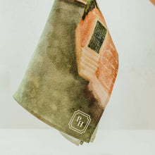 Load image into Gallery viewer, A hand holds a square kitchen towel featuring a watercolor-style print of a terracotta-colored building and green foliage. The towel has a sage green background and a small white monogram logo reading 'BH' in the bottom corner.
